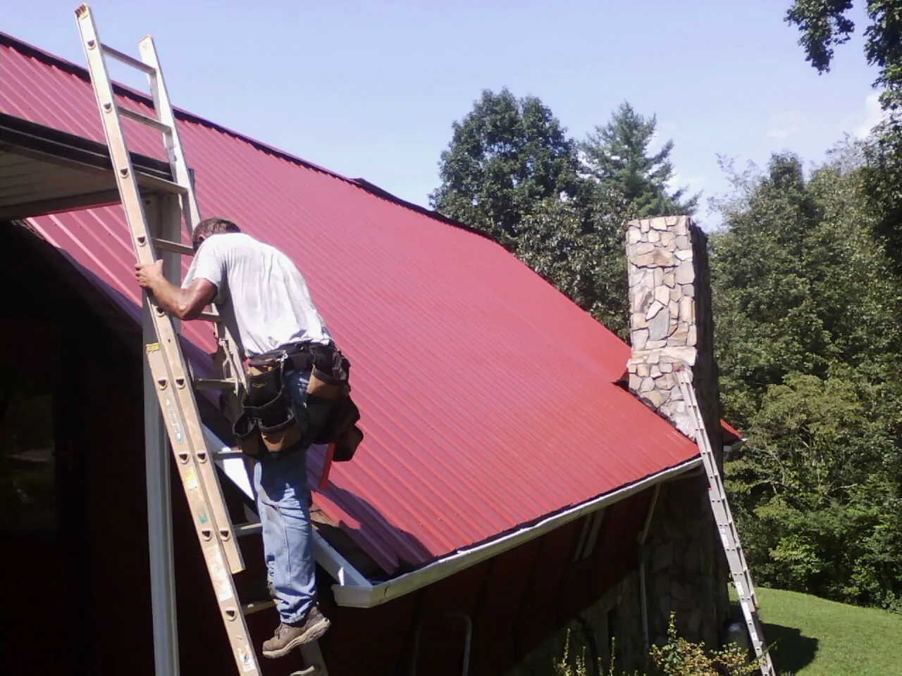 Roofer on a ladder beside a red metal roof and stone chimney — Soffit & Fascia Repair by Roofing Brothers in Temple Hills